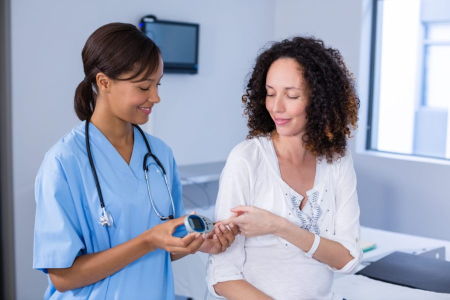 A nurse in blue scrubs measures a smiling womans blood sugar with a glucometer in a medical office. The patient sits calmly, and the nurse holds the device near her outstretched hand.