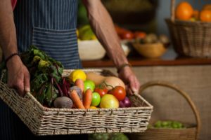 A person wearing a striped apron holds a wicker basket filled with fresh vegetables and fruits, including carrots, beets, apples, lemons, and tomatoes. Other produce baskets are visible in the background.