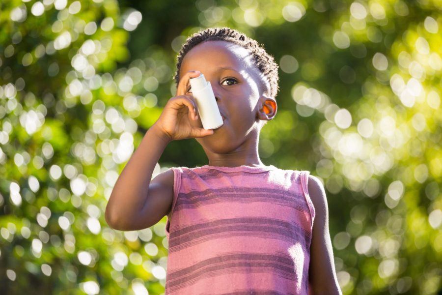 A young child in a pink striped sleeveless shirt uses an inhaler outdoors, with green foliage and sunlight blurred in the background.