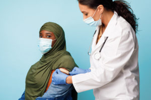 A healthcare worker in a white coat applies a bandage to the upper arm of a patient in a green hijab after vaccination. Both women wear face masks, and the background is light blue.