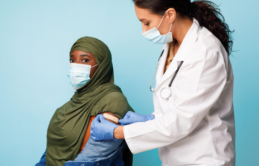 A healthcare worker in a white coat applies a bandage to the upper arm of a patient in a green hijab after vaccination. Both women wear face masks, and the background is light blue.