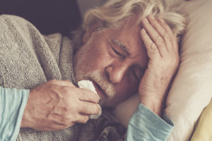 An older man with gray hair lies in bed, eyes closed, holding a tissue to his mouth and touching his forehead, appearing unwell or fatigued. He is covered with a blanket and wearing a light blue shirt.