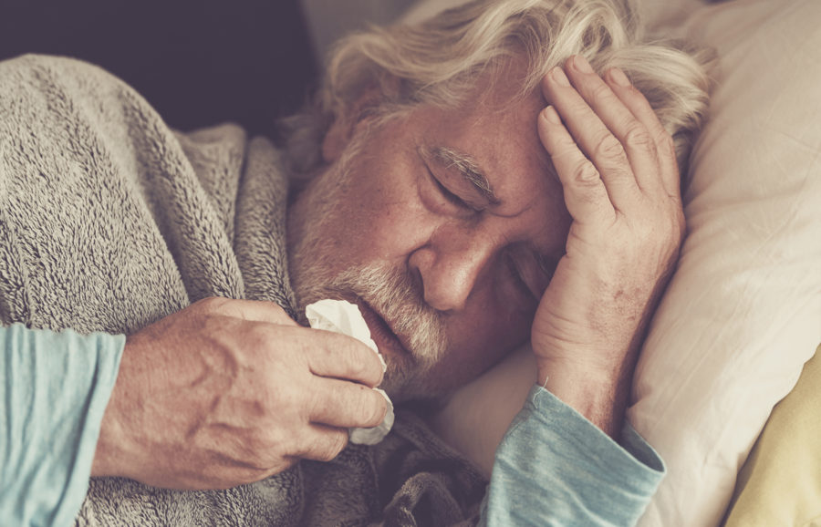 An older man with gray hair lies in bed, eyes closed, holding a tissue to his mouth and touching his forehead, appearing unwell or fatigued. He is covered with a blanket and wearing a light blue shirt.