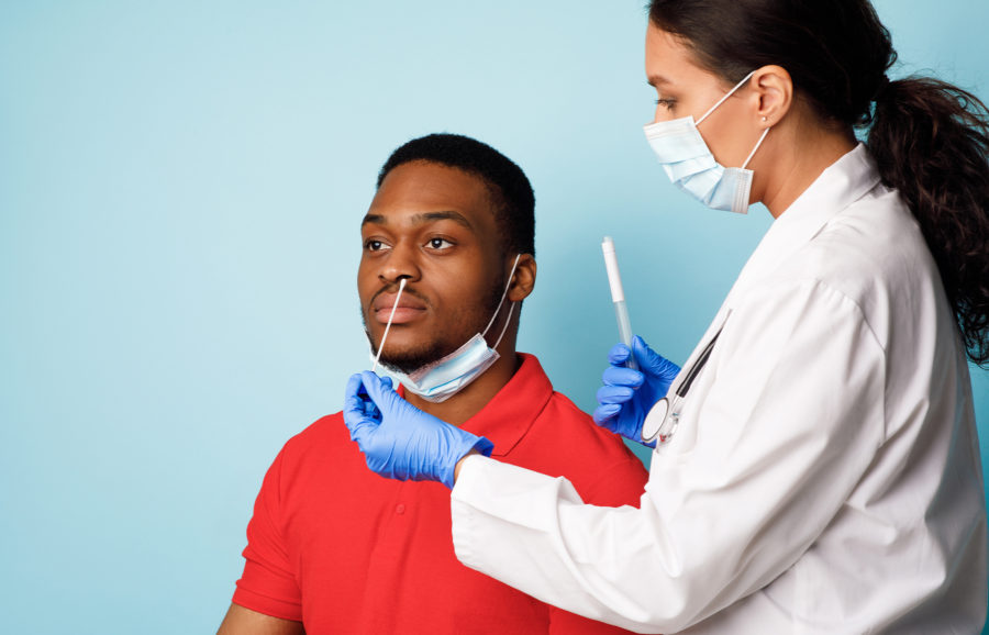 A healthcare worker wearing gloves and a mask is performing a nasal swab test on a seated man in a red shirt against a light blue background. Both are wearing surgical masks, and the man has his mask pulled down.