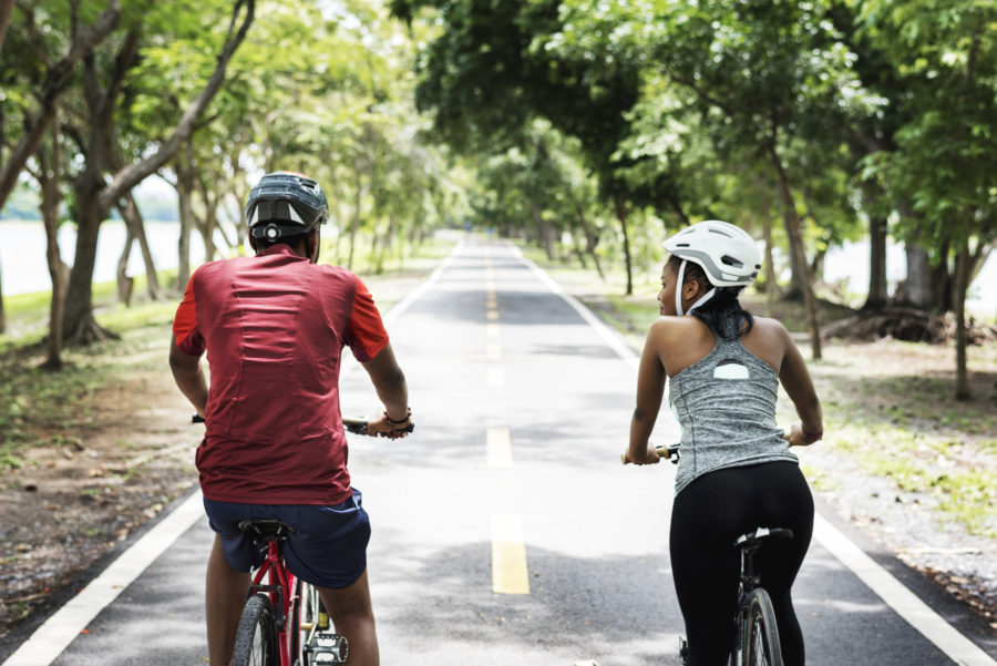 Two people wearing helmets ride bicycles side by side down a tree-lined path on a sunny day, with bright green foliage surrounding the paved trail.