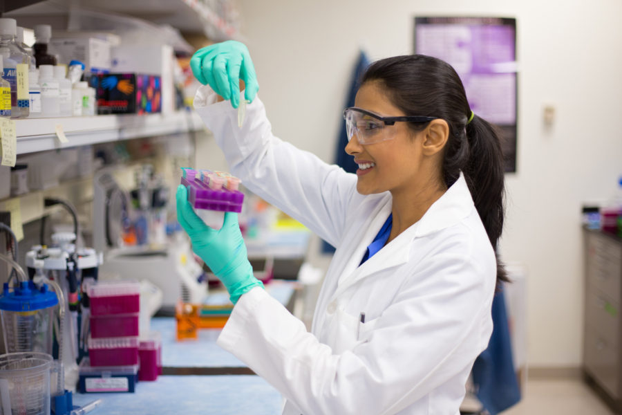 A scientist wearing a white lab coat, safety goggles, and teal gloves examines samples in a laboratory, holding a pipette and a tray of test tubes with a smile. Laboratory equipment is visible on the bench.