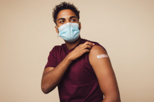 A person wearing a face mask and a maroon t-shirt shows their upper arm with a bandage, indicating they have just received a vaccination.