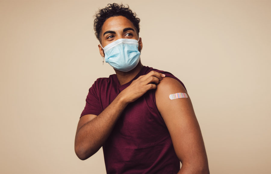 A person wearing a face mask and a maroon t-shirt shows their upper arm with a bandage, indicating they have just received a vaccination.