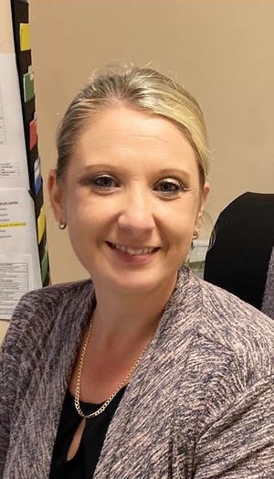 A woman with blonde hair pulled back smiles at the camera. She is wearing a patterned blazer, a black top, and a gold chain necklace. In the background, there are papers and colored file tabs on the wall.