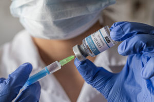 A person wearing a mask and blue gloves draws liquid from a COVID-19 vaccine vial into a syringe.