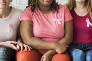 Five women sit closely together, smiling and wearing pink tops. Each has a pink ribbon pinned to their shirt, symbolizing breast cancer awareness. Their faces are partially out of frame, showing unity and support.