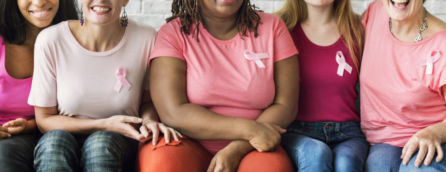 Five women sit closely together, smiling and wearing pink tops. Each has a pink ribbon pinned to their shirt, symbolizing breast cancer awareness. Their faces are partially out of frame, showing unity and support.