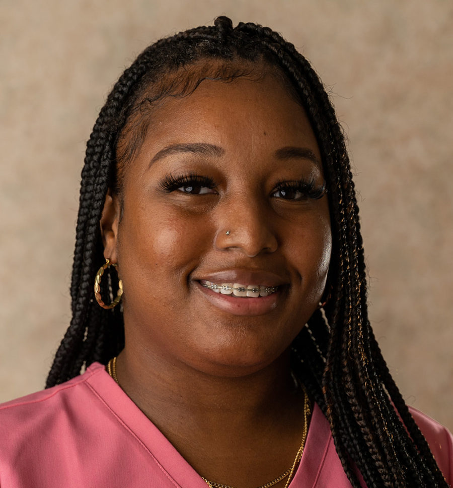 A woman with long braided hair, wearing gold hoop earrings and a pink top, smiles at the camera against a neutral background.