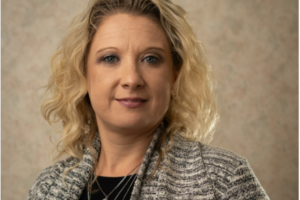 A woman with curly blonde hair wearing a gray patterned blazer and layered necklaces poses against a beige mottled background.