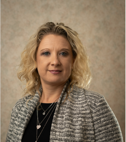 A woman with curly blonde hair wearing a gray patterned blazer and layered necklaces poses against a beige mottled background.