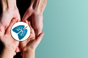 Two pairs of hands, one large and one small, gently hold a round card with a blue illustration of human lungs on a light teal background.