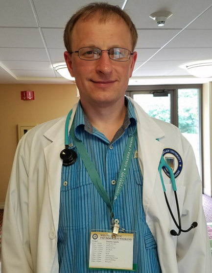 A man with glasses wearing a white lab coat and stethoscope stands indoors, smiling. He has a blue shirt and a conference name badge hanging from a lanyard around his neck.