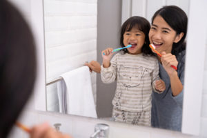 A smiling woman and a young child brush their teeth together in front of a bathroom mirror. The child wears striped pajamas with a cat design, and both look happy as they hold colorful toothbrushes.