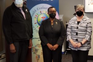 Three people wearing masks stand in front of a blue MHEDS banner in an office setting. Two women and one man are dressed in business attire. A conference table with papers is visible in the foreground.