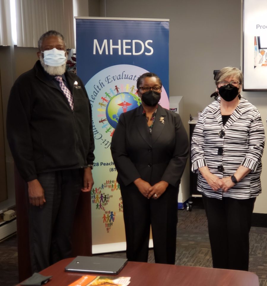 Three people wearing masks stand in front of a blue MHEDS banner in an office setting. Two women and one man are dressed in business attire. A conference table with papers is visible in the foreground.