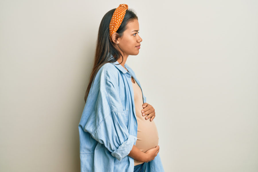 A pregnant woman in a light blue shirt and orange headband stands in profile against a plain background, gently holding her belly and looking ahead.