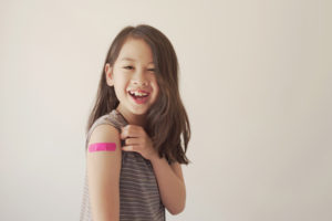 Smiling child with long hair wearing a sleeveless striped shirt shows a pink bandage on their upper arm, suggesting they recently received a vaccination, against a plain light background.