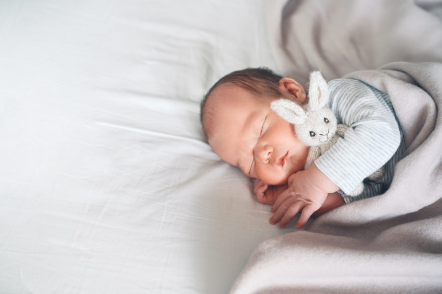 A newborn baby sleeps peacefully on a white bed, cuddling a small, white plush bunny and wrapped in a soft blanket.