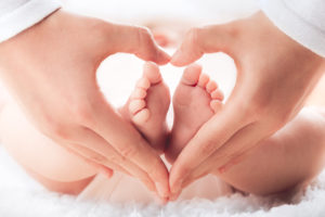 Adult hands form a heart shape around the tiny feet of a baby, who is lying on a soft, white surface. The image conveys warmth, love, and tenderness.