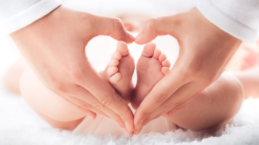 Adult hands form a heart shape around the tiny feet of a baby, who is lying on a soft, white surface. The image conveys warmth, love, and tenderness.