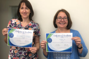 Two women smiling and holding certificates of accreditation in front of a plain white wall. Both women have glasses and shoulder-length brown hair. The certificates have blue and green borders.