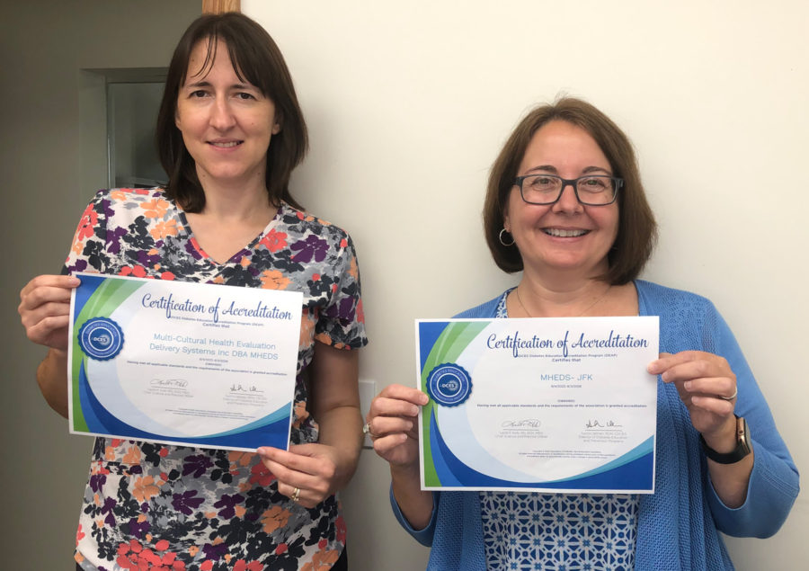 Two women smiling and holding certificates of accreditation in front of a plain white wall. Both women have glasses and shoulder-length brown hair. The certificates have blue and green borders.