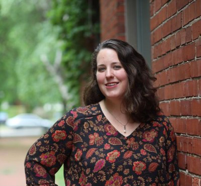 A woman with wavy dark hair, wearing a floral-patterned top, stands and smiles beside a red brick wall with greenery and blurred outdoor scenery in the background.