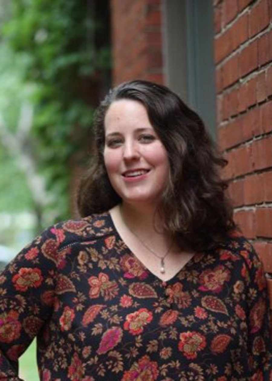 A woman with wavy brown hair wearing a black top with a colorful floral pattern stands outdoors, leaning against a red brick wall, smiling at the camera. Green foliage is visible in the background.