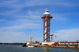 A tall observation tower with a red steel frame stands by the waters edge, near a docked sailboat, under a blue sky with scattered clouds. Trees line the distant shore.