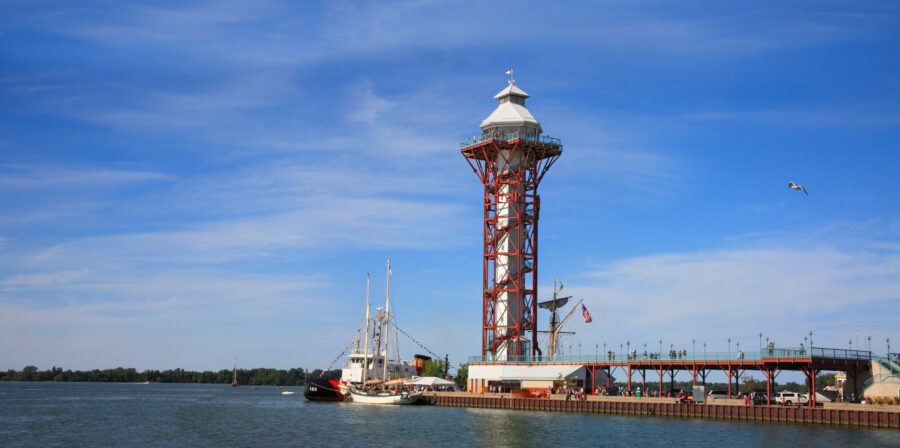 A tall observation tower with a red steel frame stands by the waters edge, near a docked sailboat, under a blue sky with scattered clouds. Trees line the distant shore.