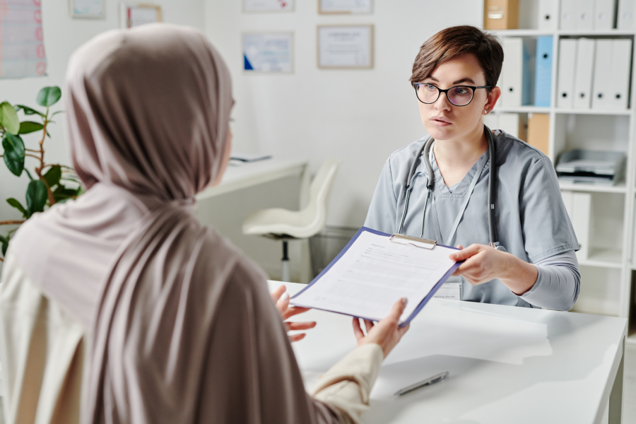 A healthcare professional in scrubs hands a clipboard with a form to a woman wearing a hijab during a consultation in a bright, modern office.