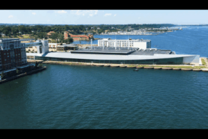 A large modern building with a sloped roof sits on the waterfront, surrounded by water on three sides. Nearby are other buildings, greenery, and boats on the bay under a partly cloudy sky.