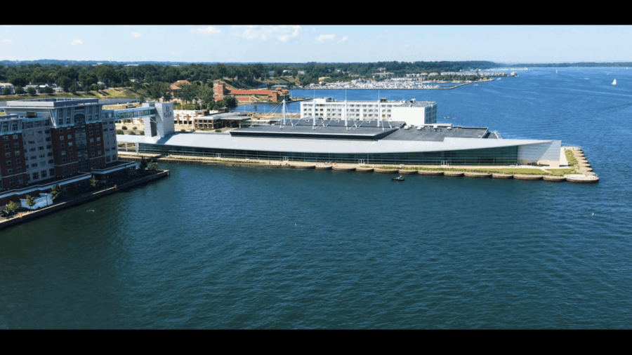 A large modern building with a sloped roof sits on the waterfront, surrounded by water on three sides. Nearby are other buildings, greenery, and boats on the bay under a partly cloudy sky.