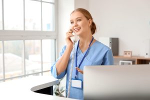 Smiling woman in blue scrubs with a name badge stands at a reception desk, talking on a corded phone in a bright office with large windows.