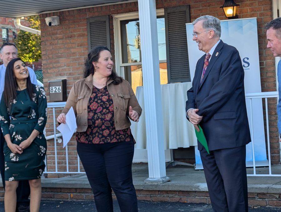 Four people stand and talk outside a brick building. A woman in a brown jacket is speaking, while others, including a man in a suit holding papers, listen and smile. A porch with white columns is in the background.