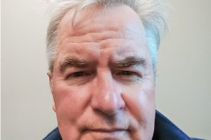 Older man with gray hair and a serious expression, wearing a blue jacket and plaid shirt, against a plain light-colored background.