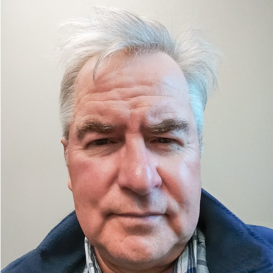 Older man with gray hair and a serious expression, wearing a blue jacket and plaid shirt, against a plain light-colored background.