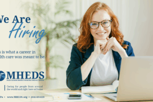 Smiling woman with red hair and glasses sits at a desk with a laptop, papers, and a pen, promoting a healthcare hiring ad for MHEDS. Text reads, We are hiring. This is what a career in health care was meant to be.