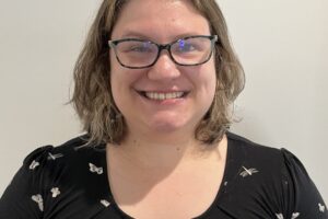 Mary Lasher smiling with shoulder-length light brown hair and glasses, wearing a black shirt with a white butterfly pattern, stands in front of a plain light-colored wall.