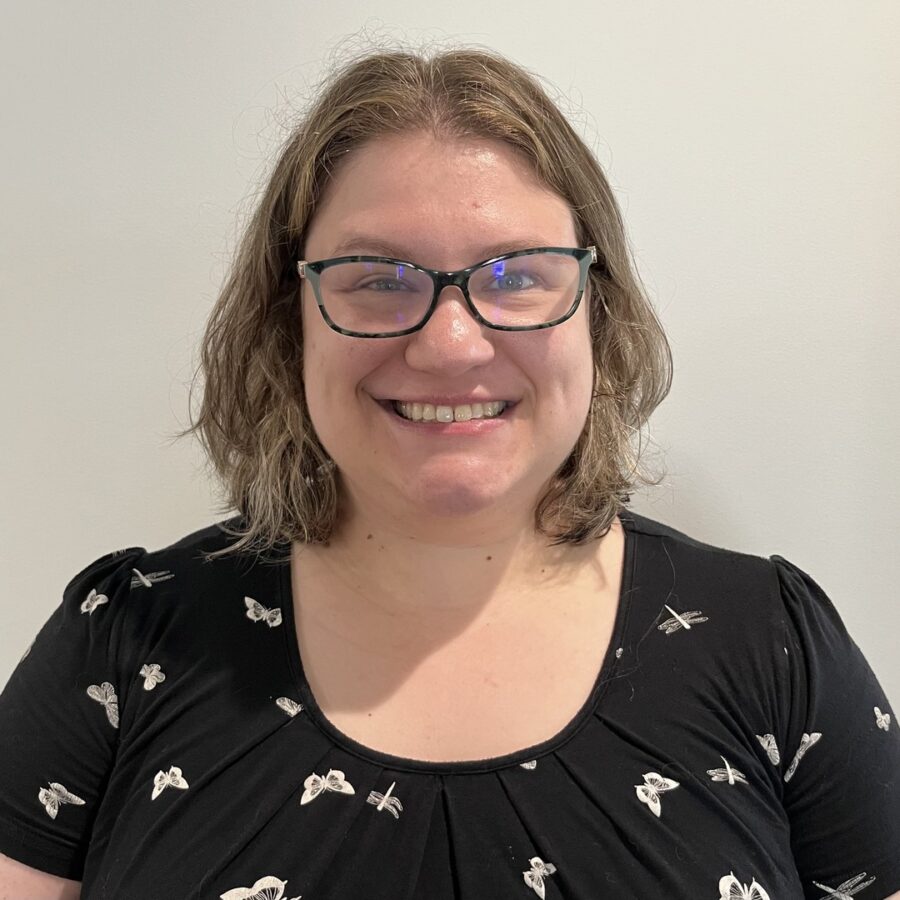 Mary Lasher smiling with shoulder-length light brown hair and glasses, wearing a black shirt with a white butterfly pattern, stands in front of a plain light-colored wall.