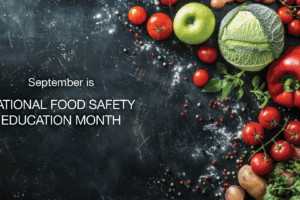 A variety of fresh vegetables and fruits, including cabbage, apples, tomatoes, peppers, and potatoes, are arranged on a dark surface with scattered spices. Text reads: September is NATIONAL FOOD SAFETY EDUCATION MONTH.