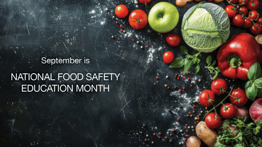 A variety of fresh vegetables and fruits, including cabbage, apples, tomatoes, peppers, and potatoes, are arranged on a dark surface with scattered spices. Text reads: September is NATIONAL FOOD SAFETY EDUCATION MONTH.