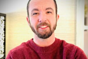 A person with short dark hair, a beard, and a nose ring is smiling at the camera. They are wearing a red sweater and are indoors with a blurred light background.