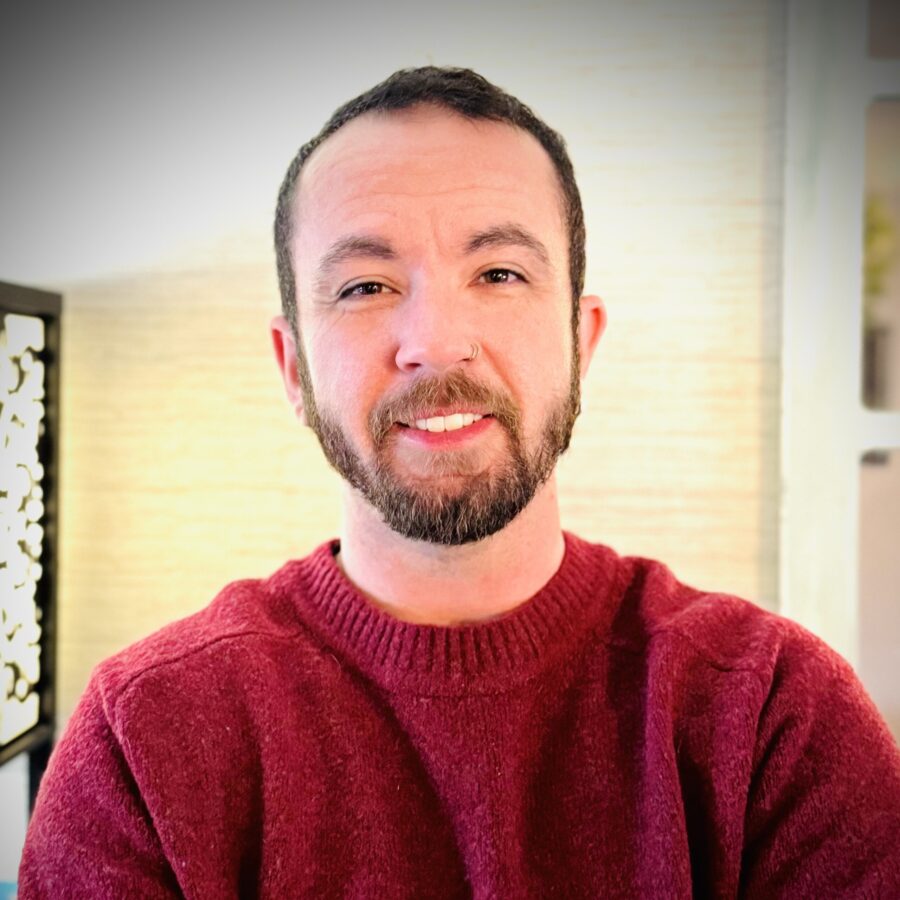 A person with short dark hair, a beard, and a nose ring is smiling at the camera. They are wearing a red sweater and are indoors with a blurred light background.