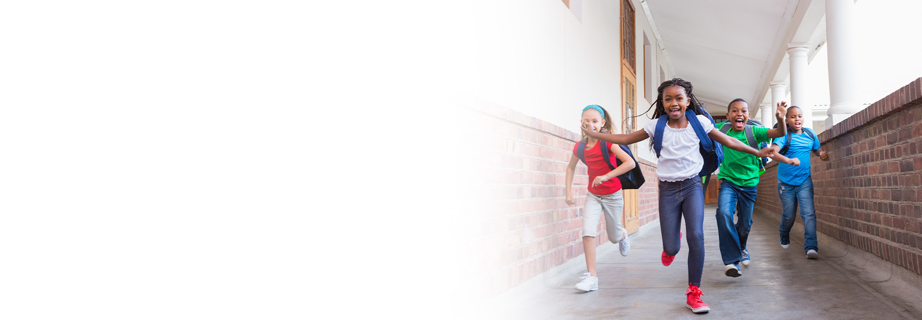 Four children with backpacks run and smile down a school hallway next to a brick wall, with bright natural light filling the scene.